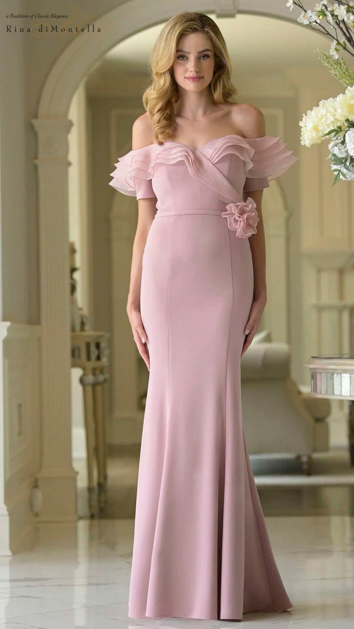 A woman wearing the Rina Di Montella RD3418 long formal dress in dusty rose—featuring a fitted sheath silhouette, off-the-shoulder neckline, ruffled sleeves, and floral waist detail—stands indoors near a white column and flowers.